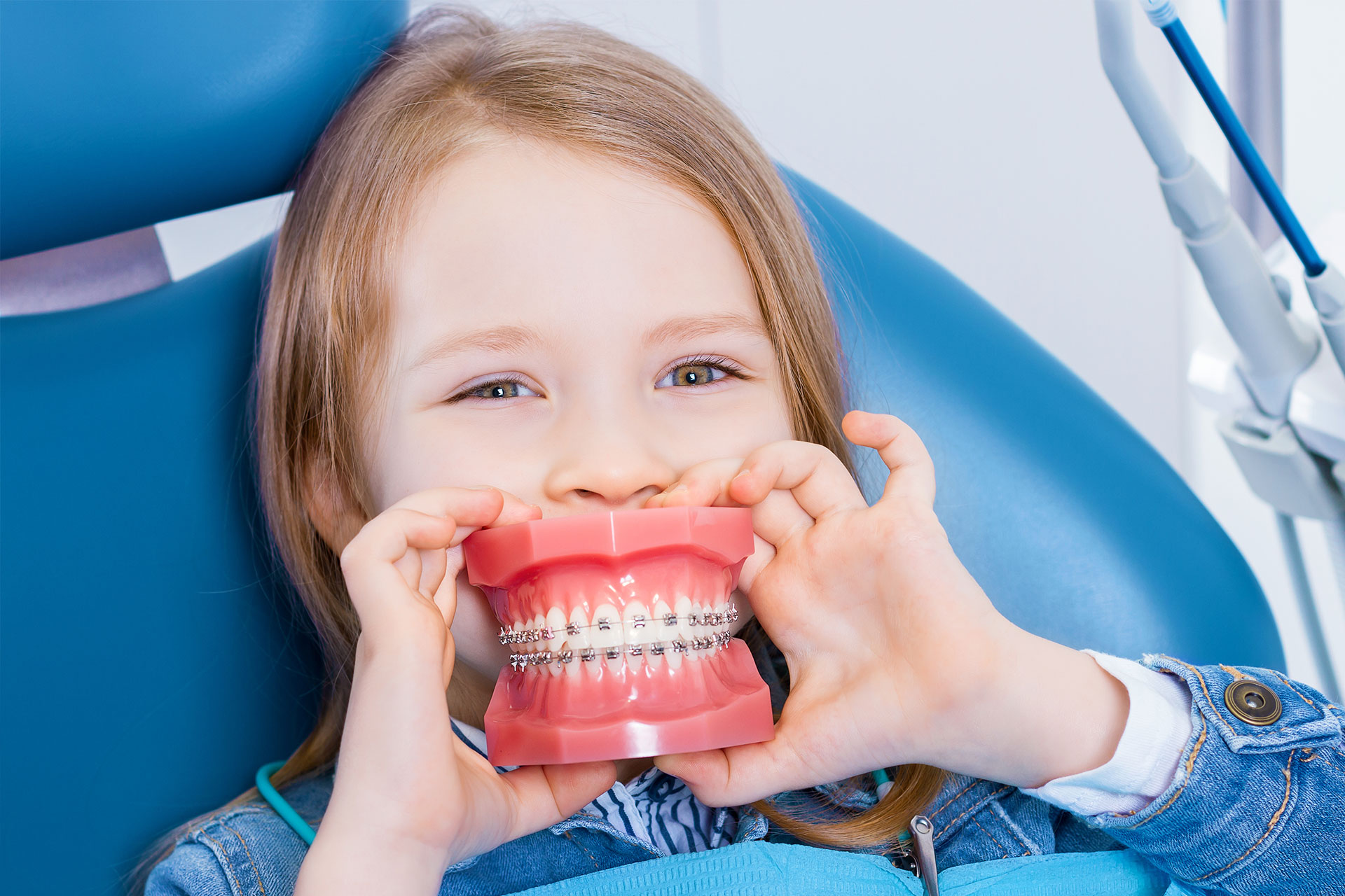 A young girl wearing braces is playfully holding her teeth open with both hands while sitting in a dental chair.