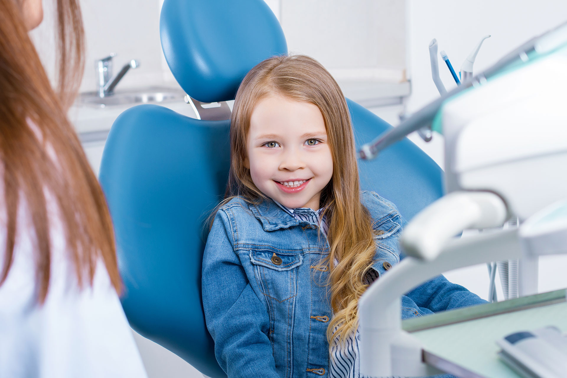 A young girl sitting in a dental chair with a dentist behind her, smiling at the camera.