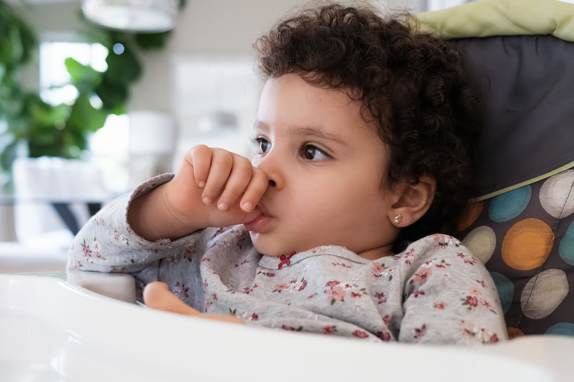 A young child with dark hair, seated in a high chair, holding their hand up to their mouth, seemingly sucking their thumb.